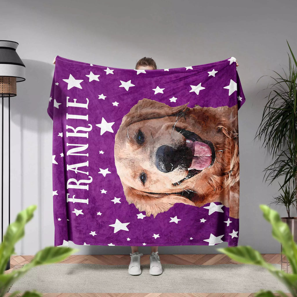 Person holding a purple pet blanket with a dog's face and 'FRANKIE' printed on it, in a room with plants and a lamp.