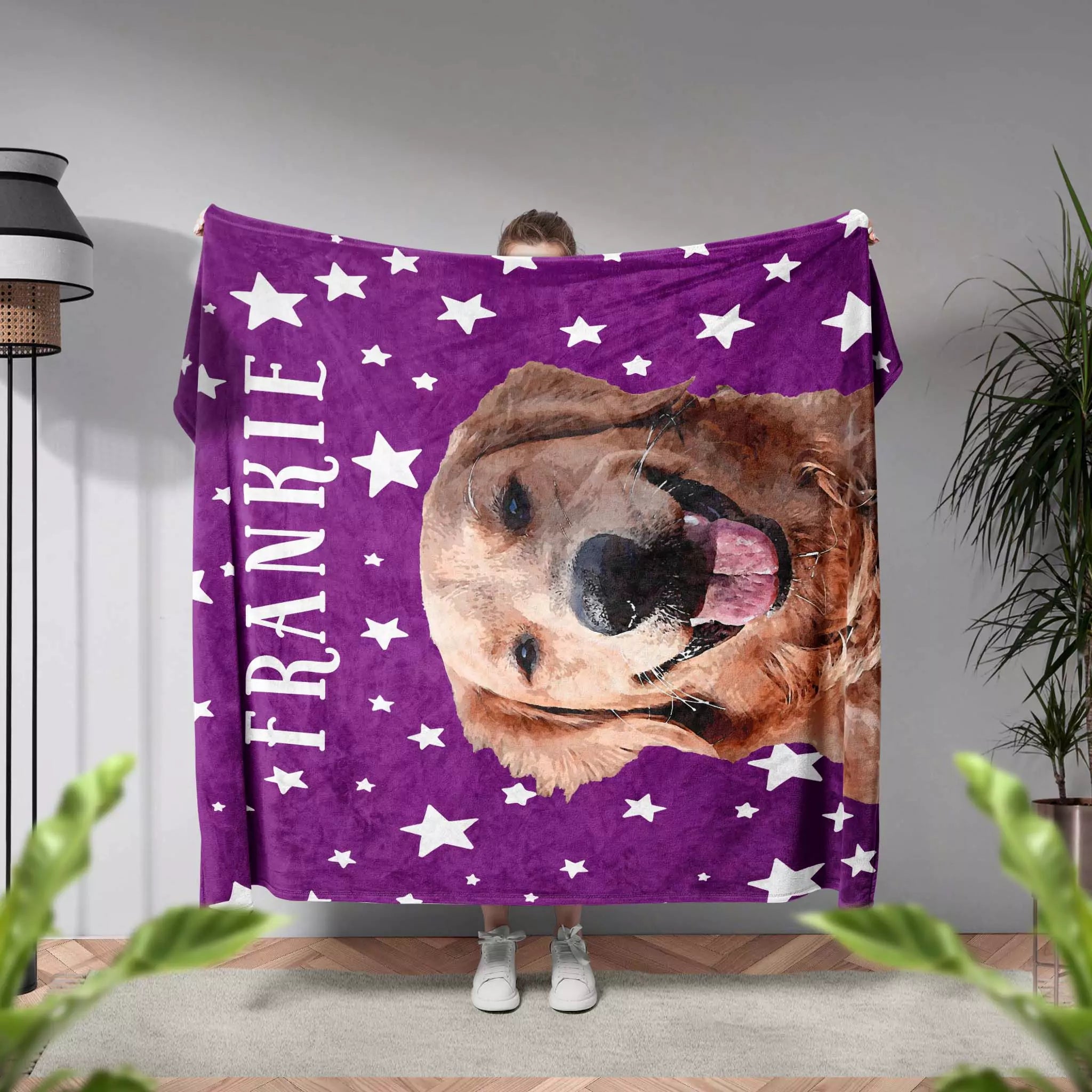 Person holding a purple pet blanket with a dog's face and 'FRANKIE' printed on it, in a room with plants and a lamp.