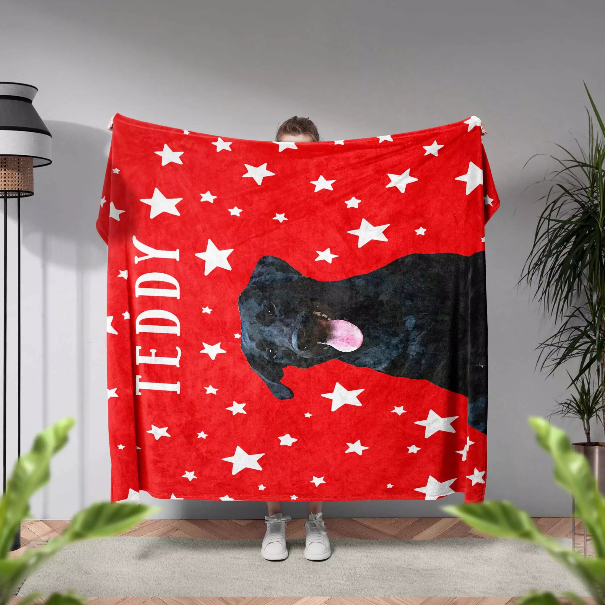 Person holding a red dog blanket with white stars and a black dog peeking out, in a room with plants and a lamp.