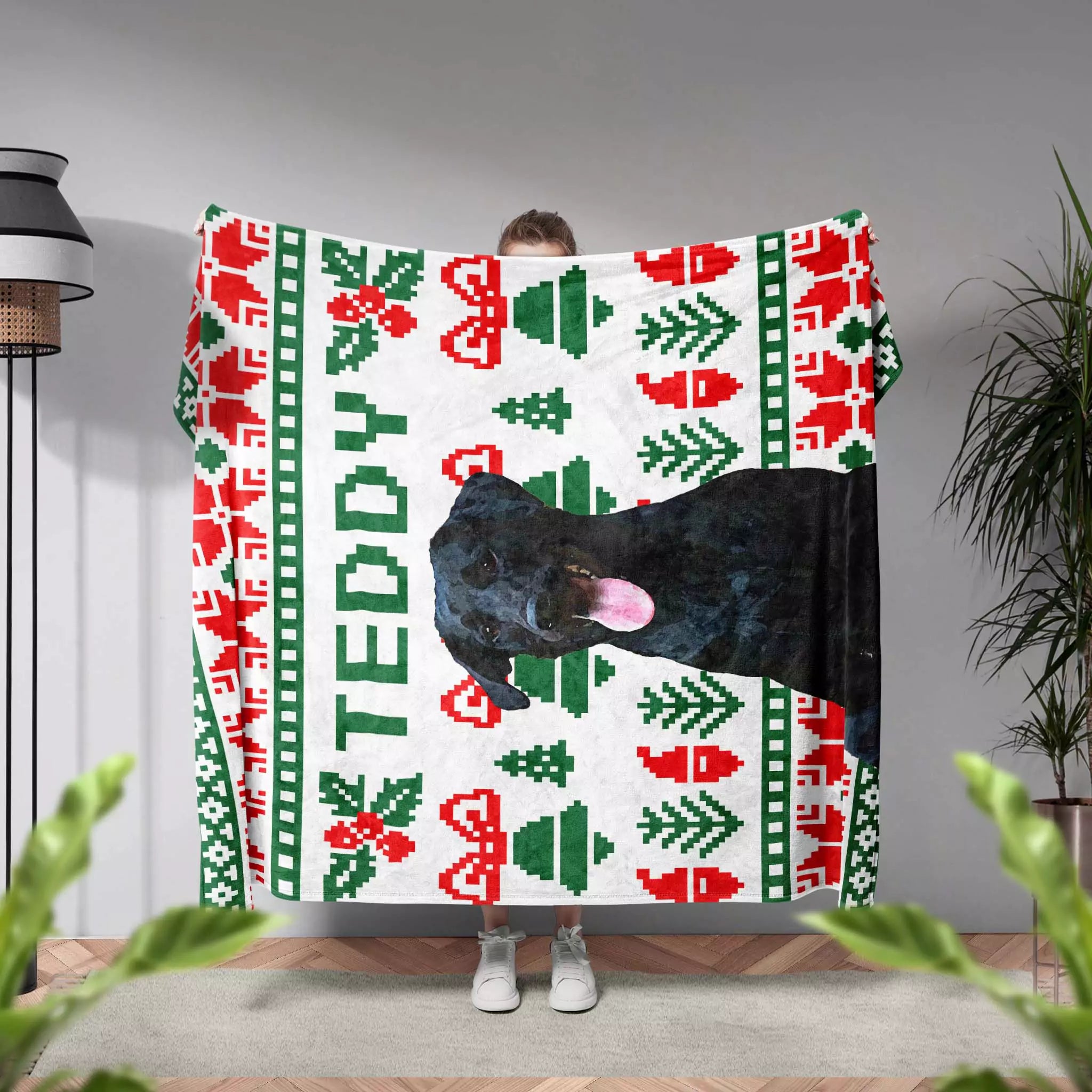 Person holding a Christmas dog blanket with a dog design and 'Teddy' text in a room with plants and a lamp.
