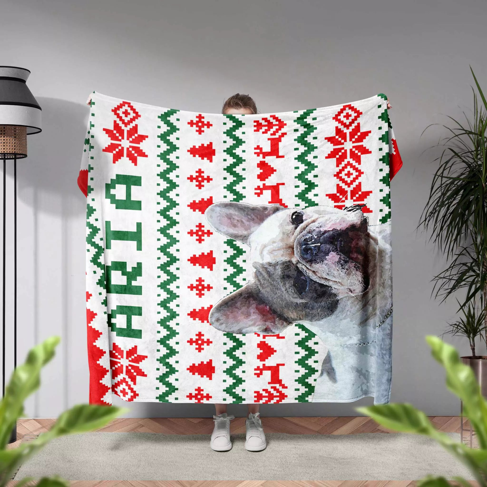 Person holding a dog blanket with a dog design and dog name text in a room with plants and a lamp.