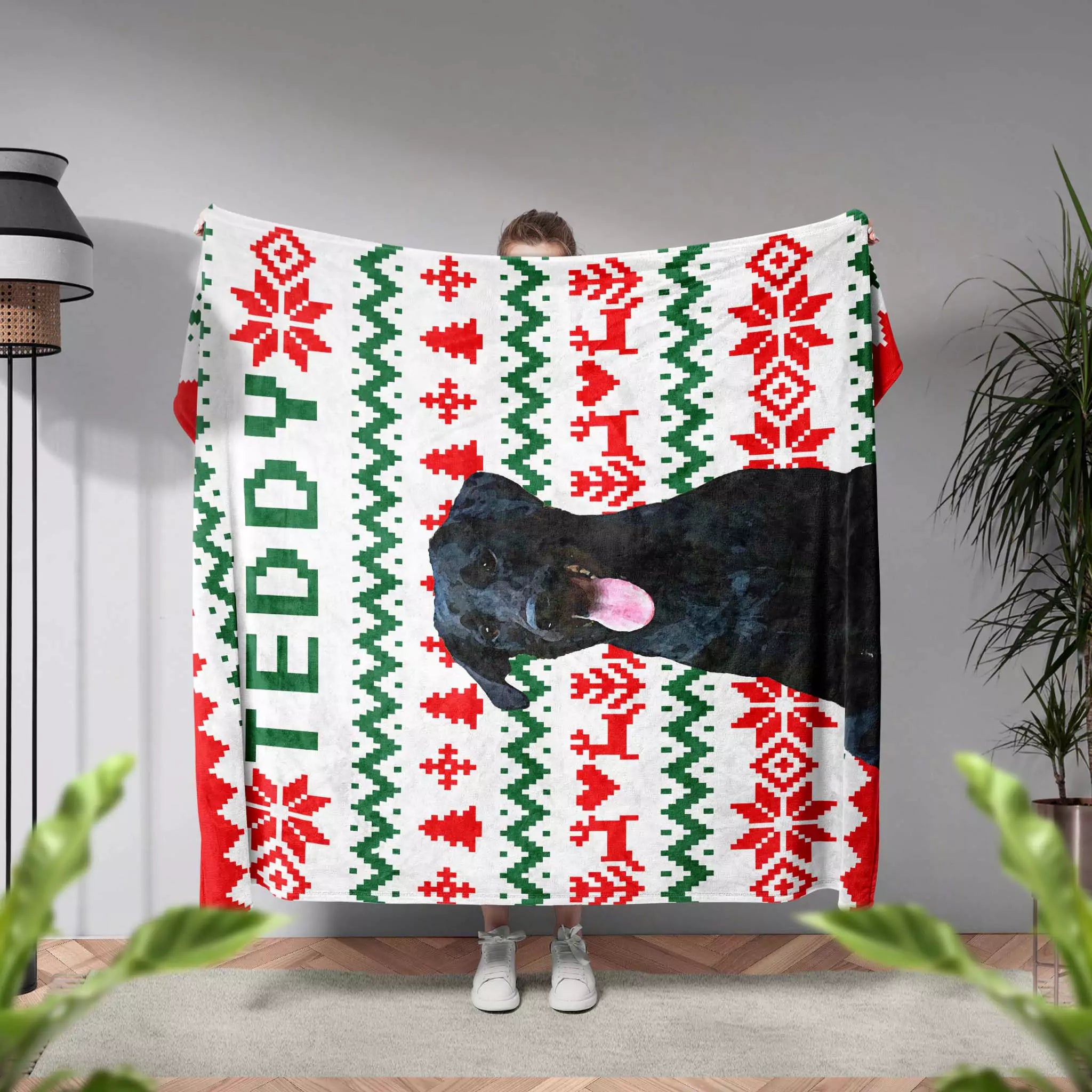 Person holding an ugly sweater blanket with a dog design and 'TEDDY' text in a room with plants and a lamp.