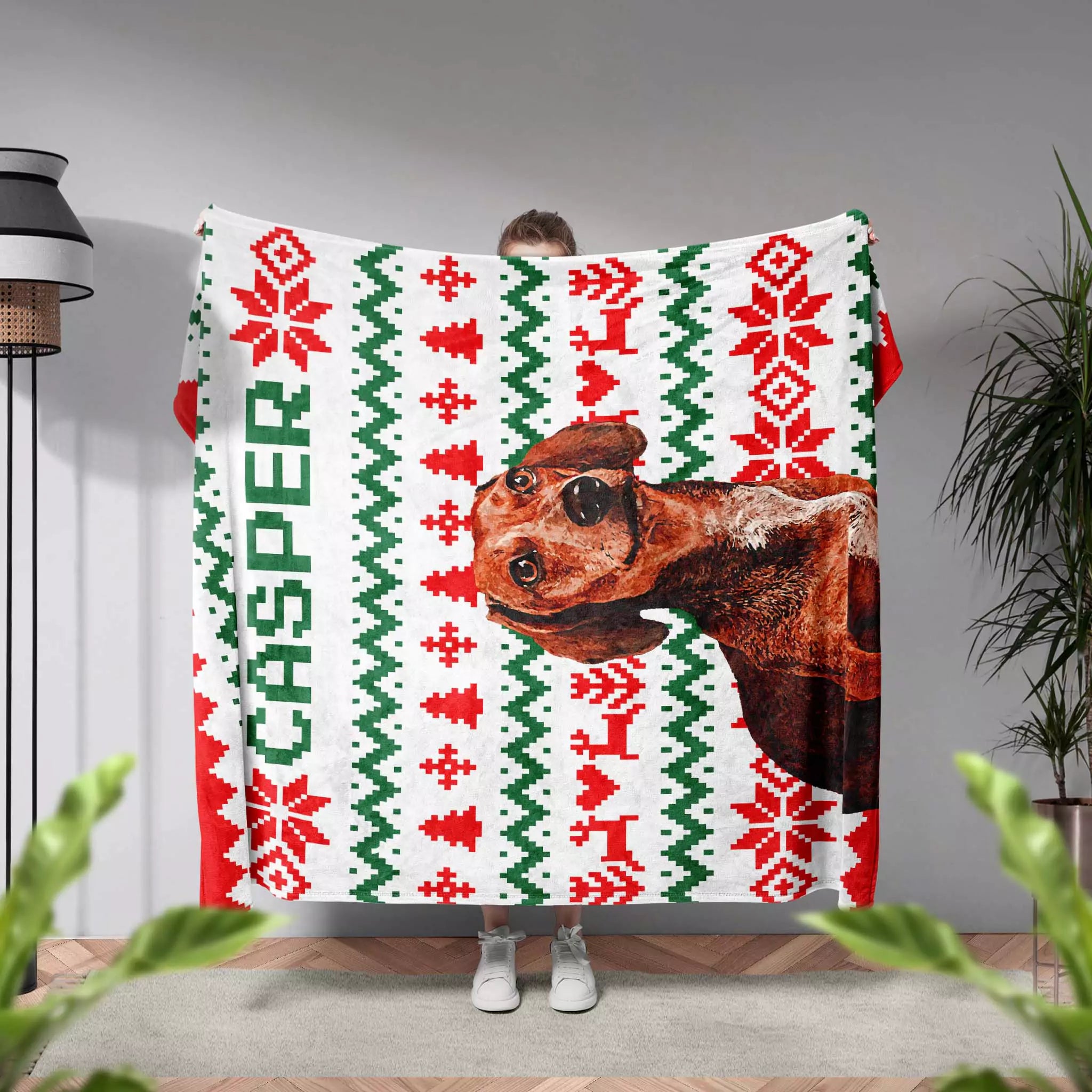 Person holding an ugly sweater blanket with a Dachshund dog design and 'Casper' text in a room with plants and a lamp.